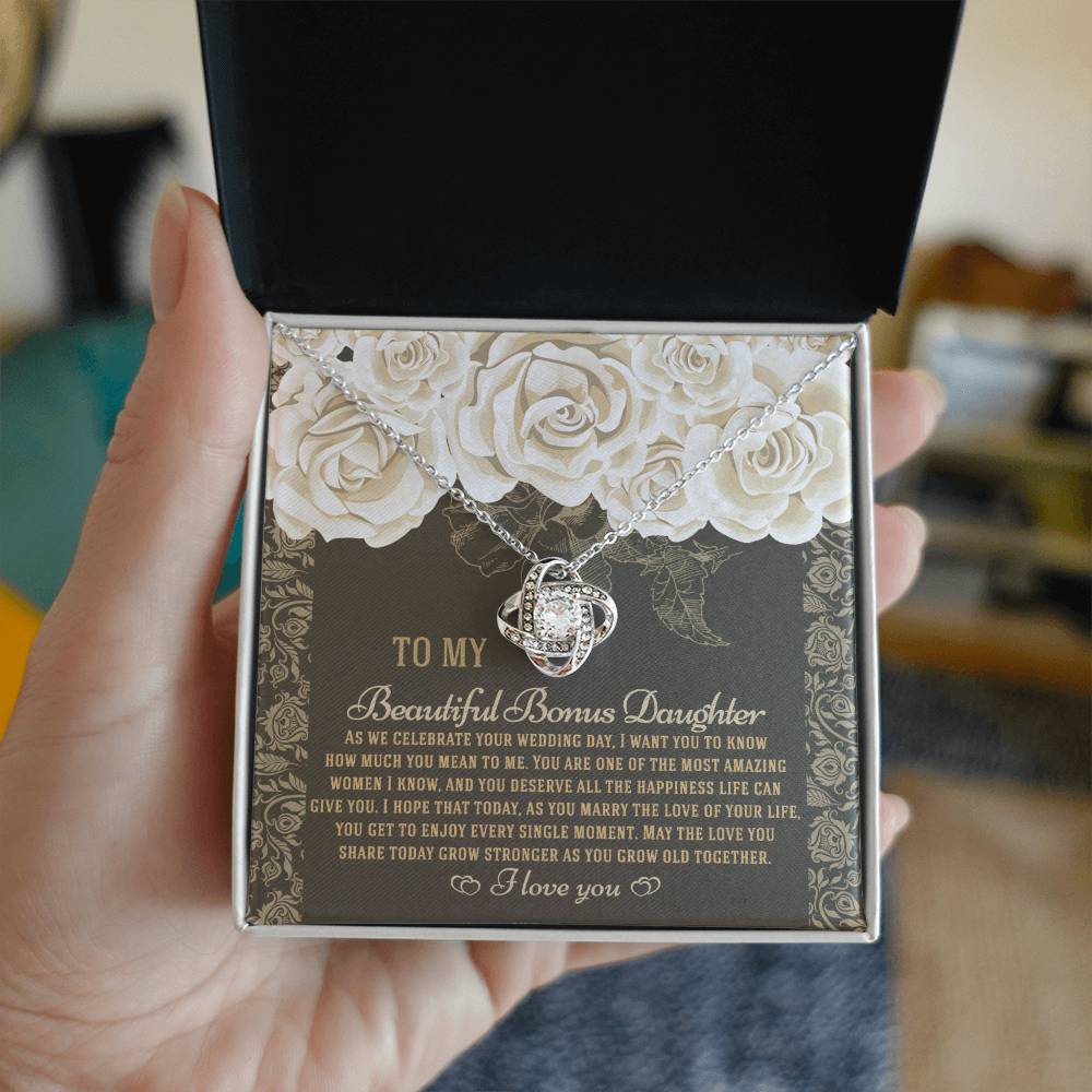 The "To Bonus Daughter, Grow Old Together - Love Knot Necklace" features a gold finish and sparkling cubic zirconia crystals. It comes packaged in a gift box decorated with an ornate floral design and includes a heartfelt message for your daughter on her wedding day.