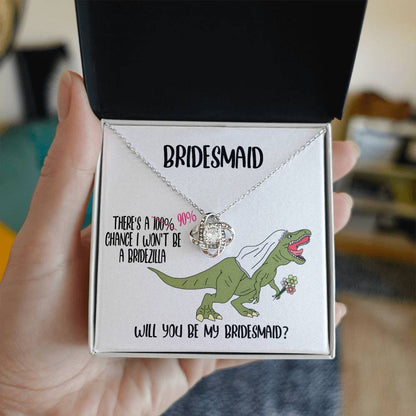 A bridesmaid proposal necklace box featuring a dinosaur with a veil and a stunning To Bridesmaid, Will You Be - Love Knot Necklace in white gold. The text reads, "There's a 90% chance I won't be a bridezilla. Will you be my bridesmaid?