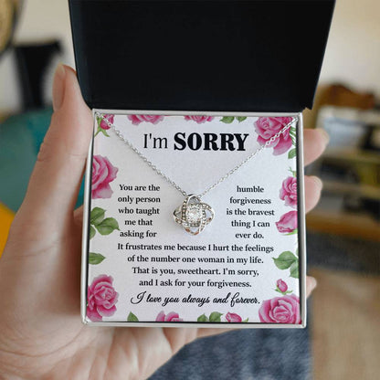 A hand holds an open box containing the "Sorry-Your Forgiveness - Love Knot Necklace," which is crafted from 14k white gold and adorned with a cubic zirconia pendant. Inside, a note that begins with "I'm Sorry" includes an apology message surrounded by pink roses.