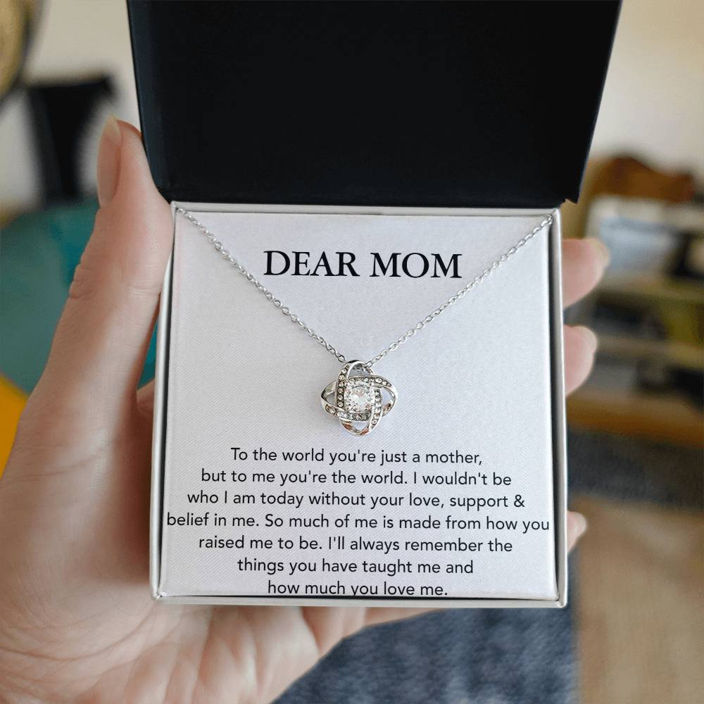 A hand holds a jewelry box containing a Dear Mom, To The World You're Just A Mother - Love Knot Necklace with an attached pendant featuring cubic zirconia crystals; inside the box lid, there is a printed message that begins with "dear mom".