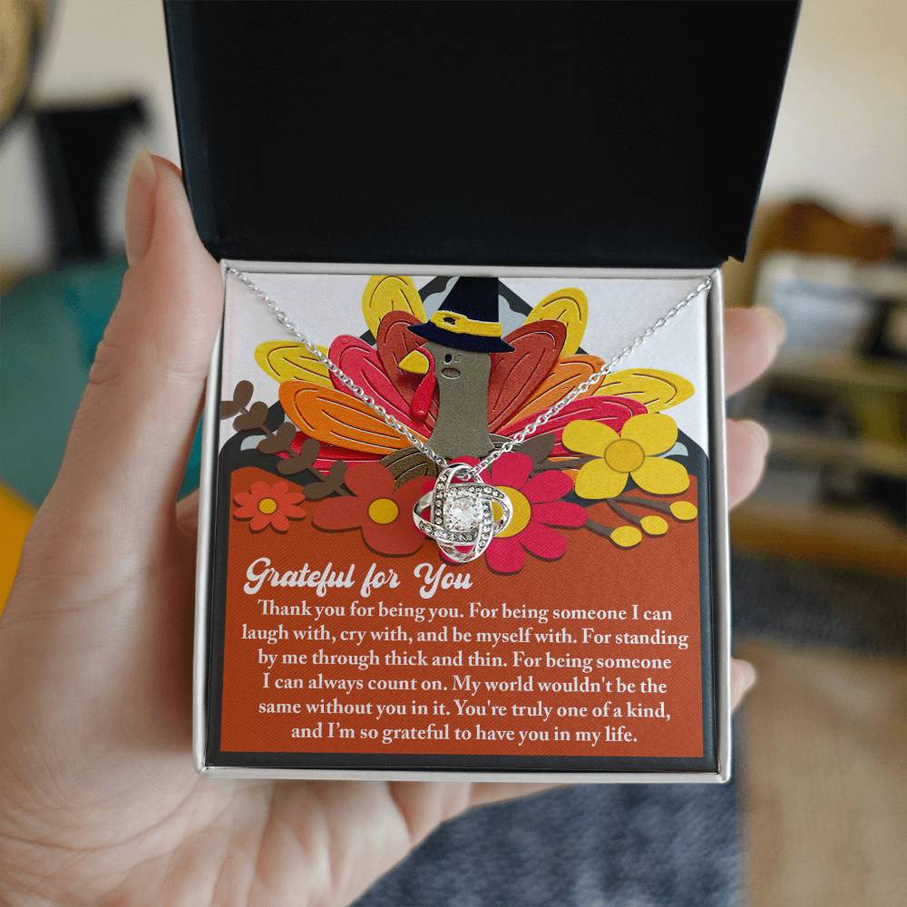 A hand holds a gift box containing the Thanksgiving-Being You - Love Knot Necklace. Inside, an illustrated card with a grateful message accompanies this beautiful necklace adorned with cubic zirconia crystals. The bright floral artwork surrounding the card adds an extra touch of personalization, making this gift truly special.