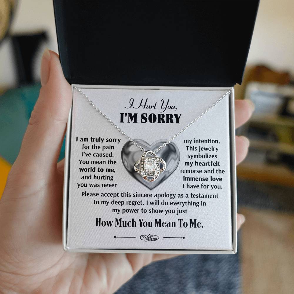 A hand holds an open jewelry box showcasing the Sorry-Sincere Apology - Love Knot Necklace, which features a heart-shaped pendant adorned with cubic zirconia crystals and includes a heartfelt message apologizing for causing pain, expressing deep regret, and promising to show immense love.