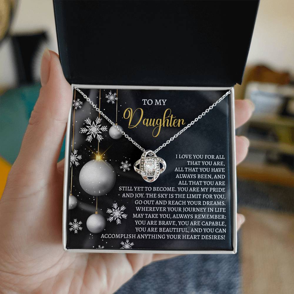 A person holds an open box containing the Daughter-Pride And Joy - Love Knot Necklace with a decorative pendant. The box lid features a heartfelt message dedicated to a daughter, making it the perfect personalized gift.