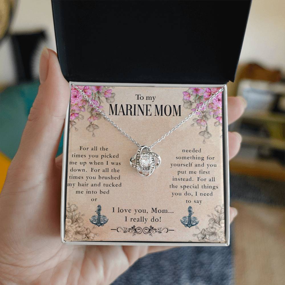 A hand is holding an open jewelry box containing a To My Marine Mom, For All The Times You Picked Me Up - Love Knot Necklace with cubic zirconia crystals and a sentimental message titled "to my marine mom," expressing appreciation and love from ShineOn Fulfillment.