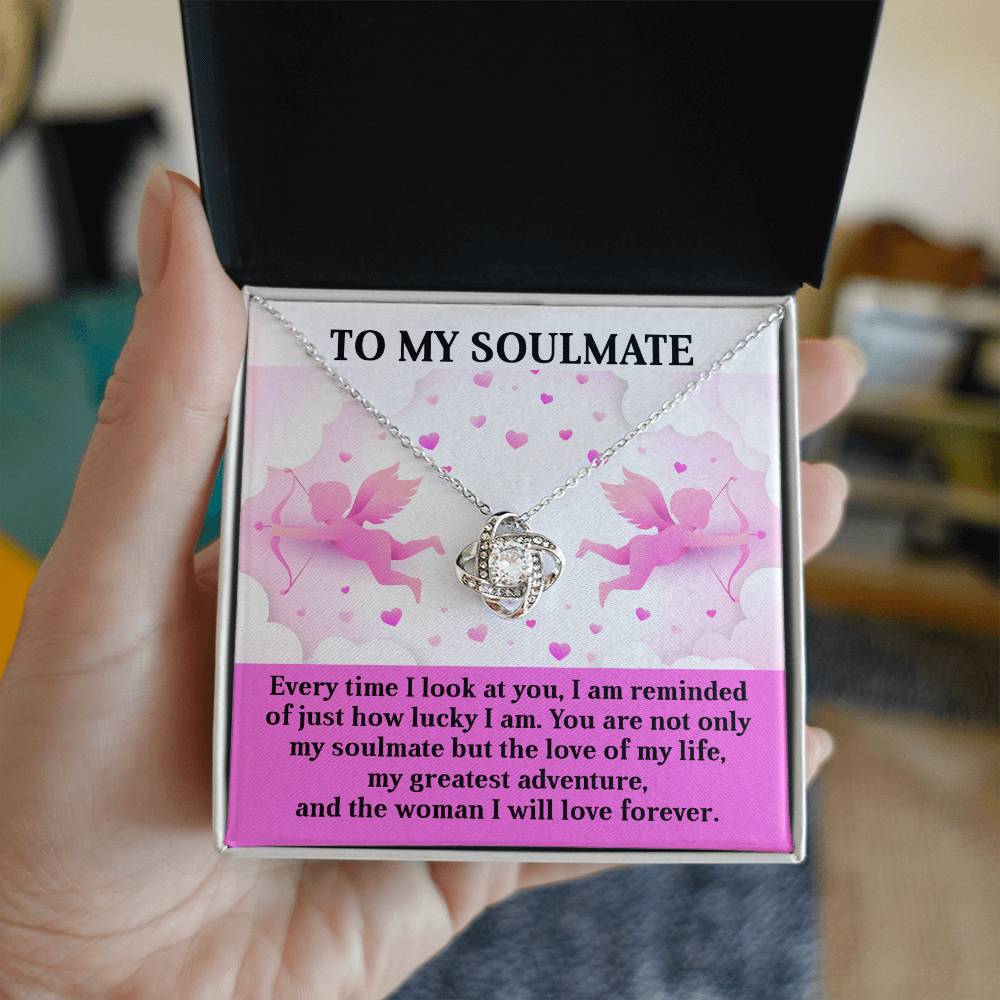 A hand holds an open jewelry box, revealing a Soulmate-Greatest Adventure Love Knot Necklace with sparkling cubic zirconia. Beside it is a card inscribed "TO MY SOULMATE" and a romantic message—an ideal personalized gift.