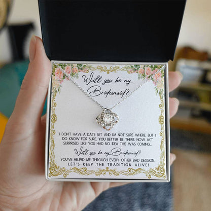 A To Bridesmaid, Keep The Tradition - Love Knot Necklace in a gift box with floral accents, featuring 14k white gold and cubic zirconia crystals. The message reads: "Will you be my Bridesmaid? I don't have a date set... act surprised. You've helped me through every other bad decision. Let's keep the tradition alive!