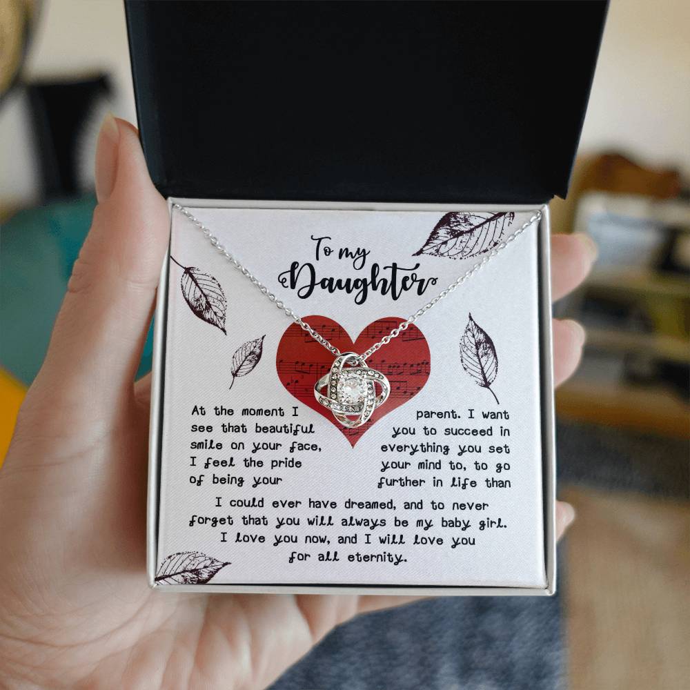 A person holds an open gift box containing a Daughter-On Your Face - Love Knot Necklace, adorned with cubic zirconia crystals, and a card with a heartfelt message addressed "To my Daughter," expressing love and encouragement.