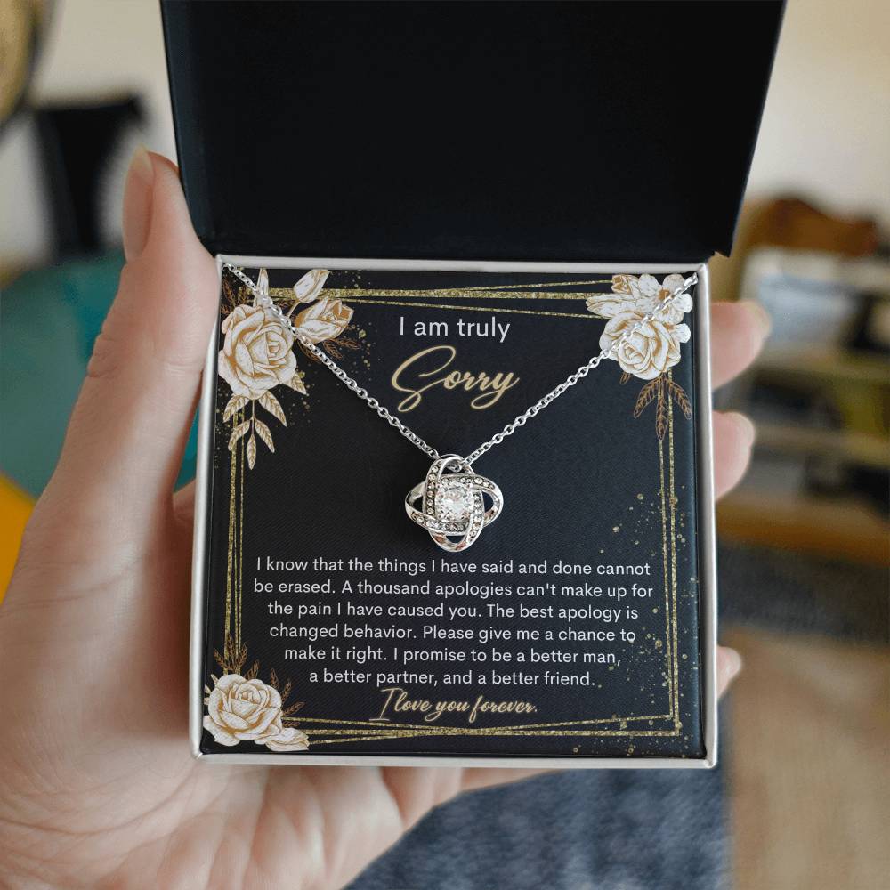 The Sorry-Cannot Be Erased - Love Knot Necklace, adorned with shimmering cubic zirconia crystals and available in a choice of white gold or yellow gold finish, is displayed against a backdrop of a black card bearing an apology message that reads, "I am truly sorry" and "I promise to be a better man, a better partner, and a better friend.