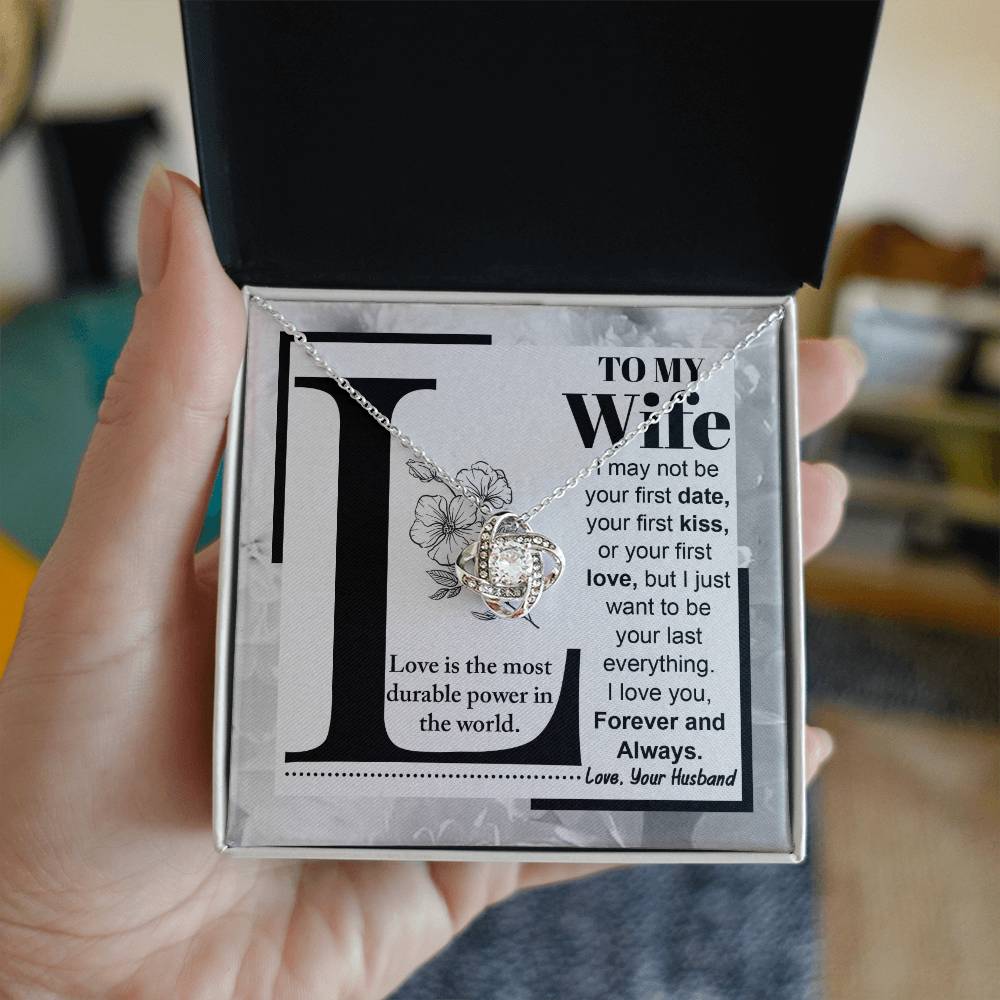 A "to Wife, Love Is - Love Knot Necklace" with a knot design is displayed on a card featuring a printed message to a wife about lasting love, signed from a husband. The piece sparkles with cubic zirconia crystals and boasts an elegant gold finish.