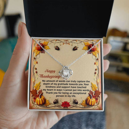 A hand holds an open jewelry box containing the Thanksgiving-Put Into Words - Love Knot Necklace, embellished with sparkling cubic zirconia crystals. The box features a Thanksgiving-themed message adorned with autumn leaves and pumpkins, making it an ideal personalized gift for the season.