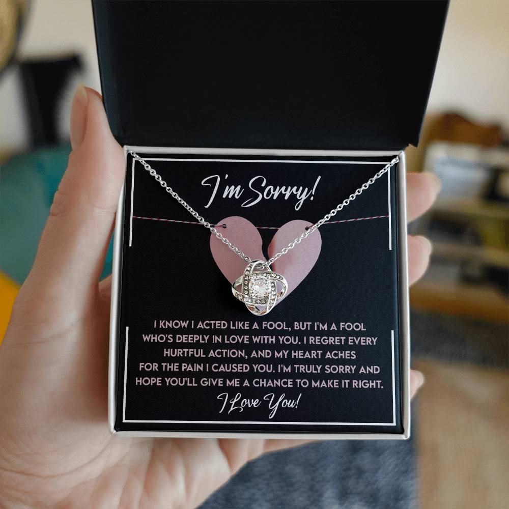 A hand holds an open box with the Sorry-Deeply In Love - Love Knot Necklace inside, adorned with premium cubic zirconia crystals. The box has a heartfelt apology message written on the inner lid, expressing regret and love—making it a perfect personalized gift.