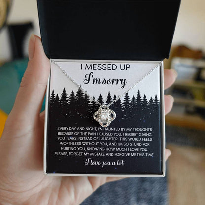 The Sorry-By My Thoughts - Love Knot Necklace, featuring a heart-shaped pendant adorned with cubic zirconia crystals, is displayed on a card with an apology message. The card reads: "I messed up. I'm sorry. Every day and night, I'm haunted...I love you a lot," set against a serene forest background.