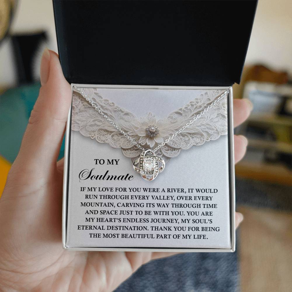 A hand holds an open box containing the Soulmate-Eternal Destination - Love Knot Necklace, crafted with premium cubic zirconia. The box cover features a heartfelt message about love and soulmates, making it a perfect personalized gift.