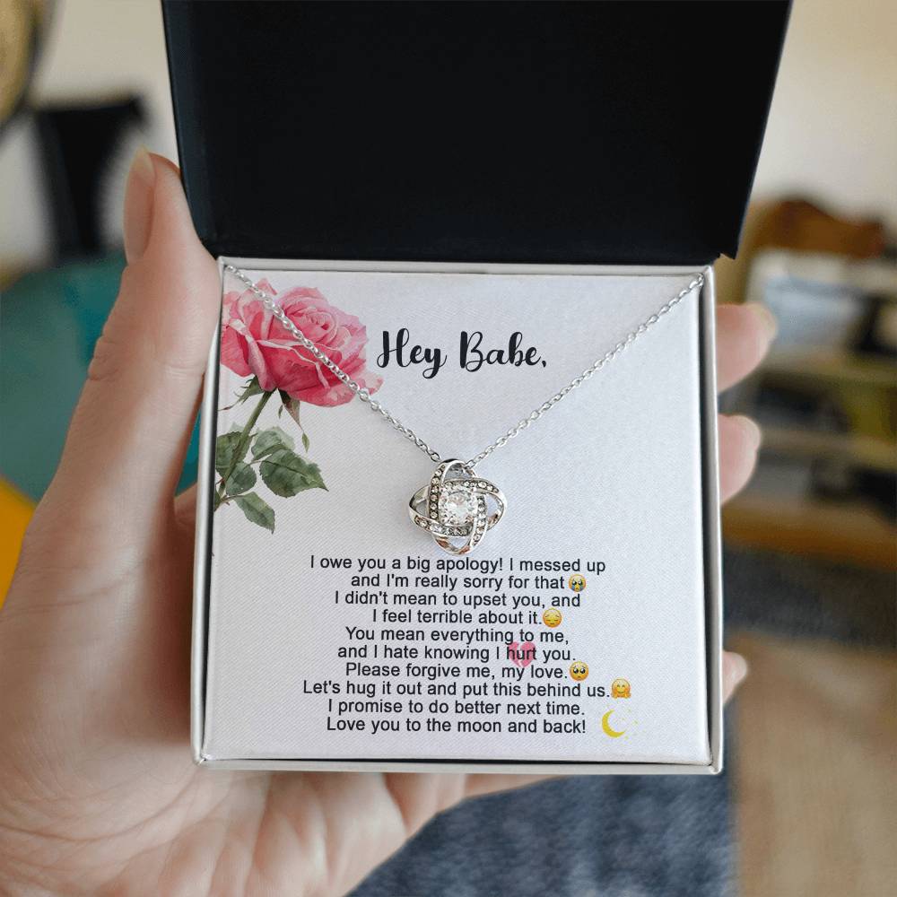 The "Sorry-Hug It Out - Love Knot Necklace" features an 18k yellow gold pendant adorned with small cubic zirconia crystals. It comes on a card decorated with roses, accompanied by the message "Hey Babe" and an apology note.