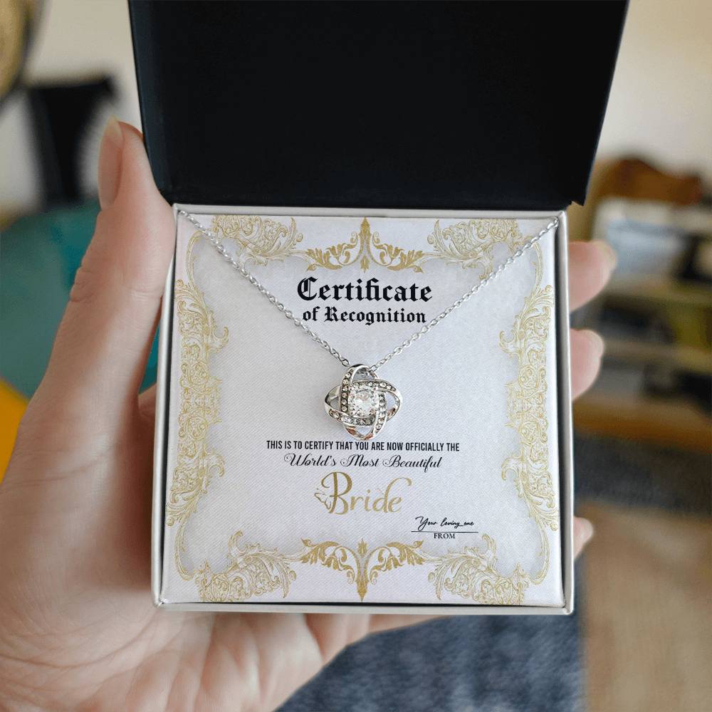 A To Bride, Certificate Of Achievement - Love Knot Necklace with a diamond-studded pendant is inside a box labeled "Certificate of Recognition" with text declaring "World's Most Beautiful Bride" and a space for names "You bring me" and "From." The pendant, set in 14k white gold, sparkles brilliantly.