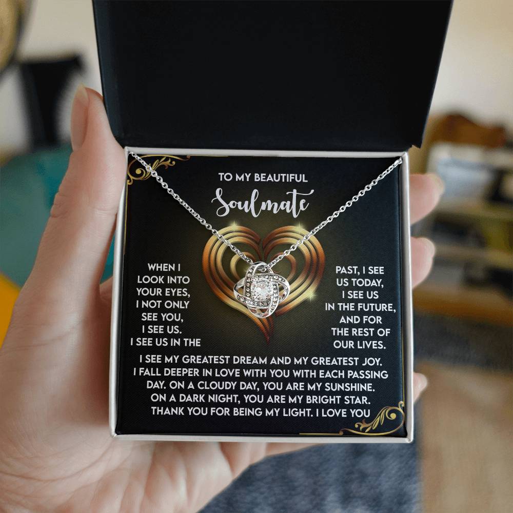 A person unveils an open gift box containing the "Soulmate-I See Us - Love Knot Necklace," sparkling with cubic zirconia crystals. The box features a heart and floral design with a romantic message to a soulmate, ideal for a personalized gift.