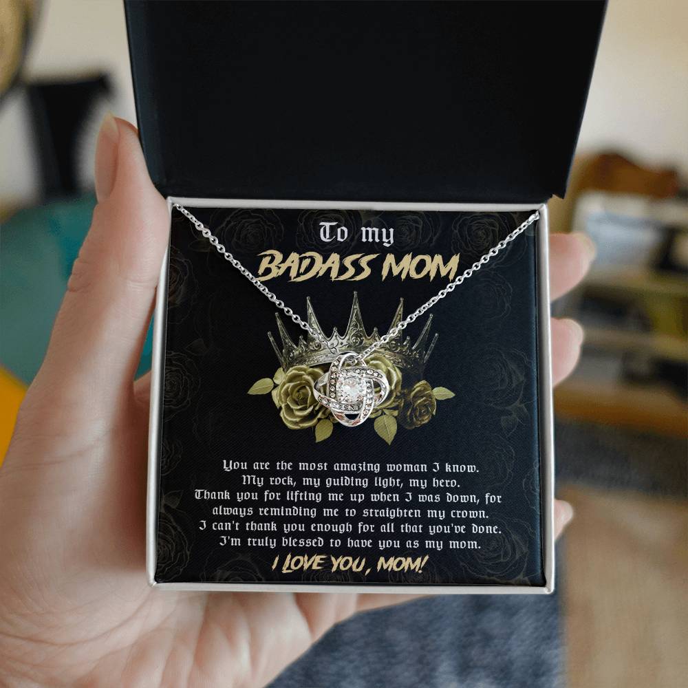 A person holds an open box containing a silver "To Mom, Straighten My Crown - Love Knot Necklace" adorned with cubic zirconia crystals, featuring a crown and rose design. The box lid reads "To my BADASS MOM" and a heartfelt message to the mother is printed inside, making it the perfect personalized gift.