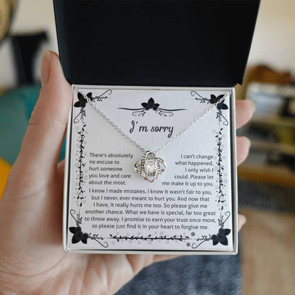 The Sorry-No Excuse - Love Knot Necklace, with a white gold finish, showcases a delicate knot design on a chain adorned with cubic zirconia crystals. The pendant is presented on a box featuring an "I'm sorry" message that expresses regret and seeks forgiveness, framed by a charming floral border at the top.