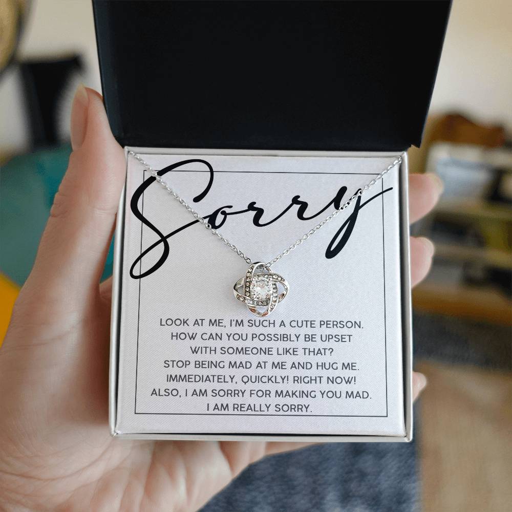 A hand holds an open box with the Sorry-Someone Like That - Love Knot Necklace adorned with cubic zirconia crystals and a message that reads, "Sorry. Look at me, I'm such a cute person... I am really sorry.