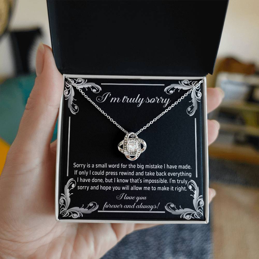 The "Sorry, Make It Right 2 - Love Knot Necklace" showcases a silver interlinked design on a black background. The backdrop features an apology message written in white text, complemented by the brilliance of cubic zirconia crystals.