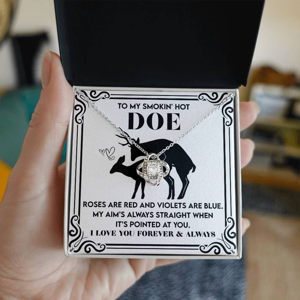 A hand holds an open box containing a Gold Finish Love Knot Necklace from the "Soulmate-Pointed At You" collection. The box reads, "To my smokin' hot doe," with a romantic message, and features a deer illustration with sparkling Cubic Zirconia heart on an enchanting background.