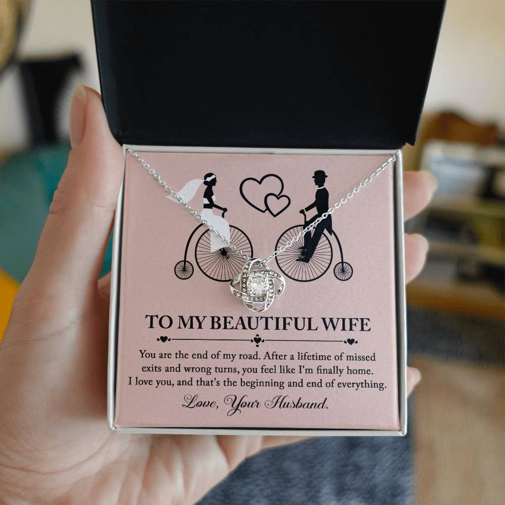 To Wife, I'm Finally Home - Love Knot Necklace with intertwined circle pendant on a card featuring an illustration of a couple on bicycles. The necklace, adorned with cubic zirconia crystals and a stunning gold finish, comes with a card that reads: 'To my beautiful wife' followed by a romantic message and ending with 'Love, Your Husband.'