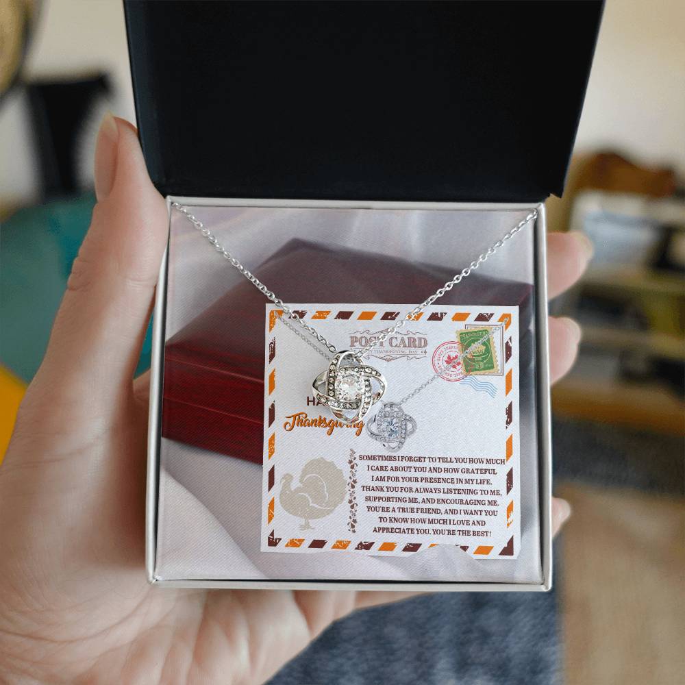 An open jewelry box, showcasing the "Thanksgiving-A True Friend - Love Knot Necklace" embellished with cubic zirconia crystals, is held in a hand against a postcard backdrop featuring decorative stamps and text.