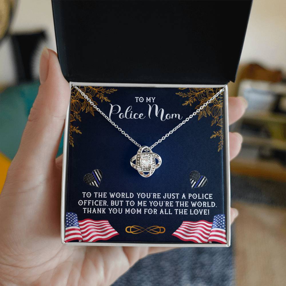 Sentence with replaced product: A hand holding an open jewelry box containing a To My Police Mom, To The World You're Just A Police Officer - Love Knot Necklace pendant, dedicated to a 'police mom,' with an affectionate message of appreciation printed inside the lid from ShineOn Fulfillment.