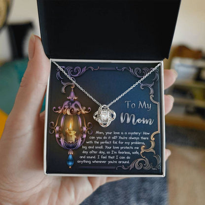 A hand holds an open box containing the "To Mom, Love Is Mystery - Love Knot Necklace," showcasing a globe-shaped pendant. The background features a decorative card with text expressing love and appreciation to a mom. The pendant's intricate design is highlighted by its gold finish and sparkling cubic zirconia accents.