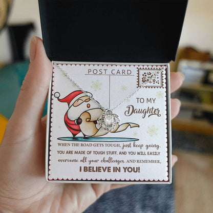 A hand presents an open jewelry box showcasing the Daughter-Keep Going - Love Knot Necklace, embellished with shimmering cubic zirconia. The accompanying card reads "To my Daughter," featuring an encouraging message and a Santa illustration, symbolizing your unbreakable bond.