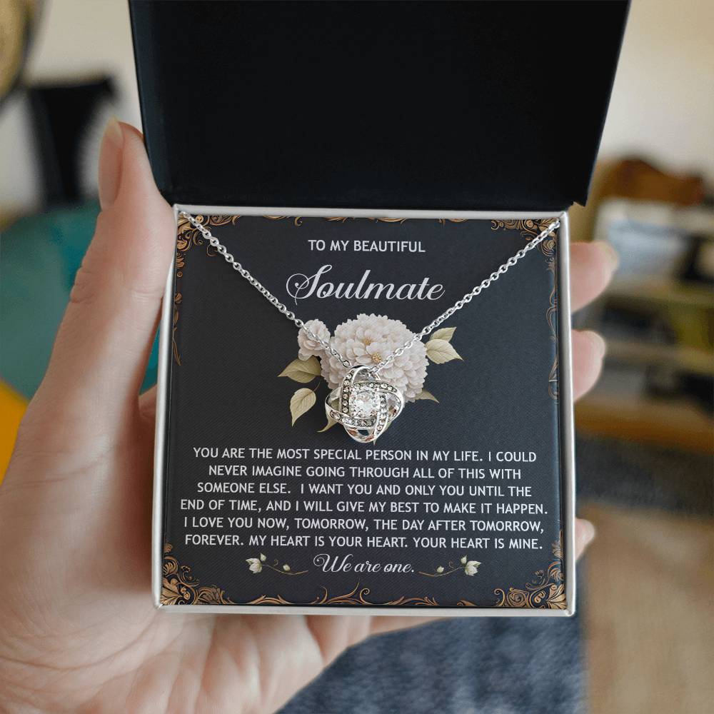 A hand holds an open box with a Soulmate-We Are One Love Knot Necklace featuring cubic zirconia crystals. The box interior displays a heartfelt message to a soulmate, making it the perfect personalized gift.