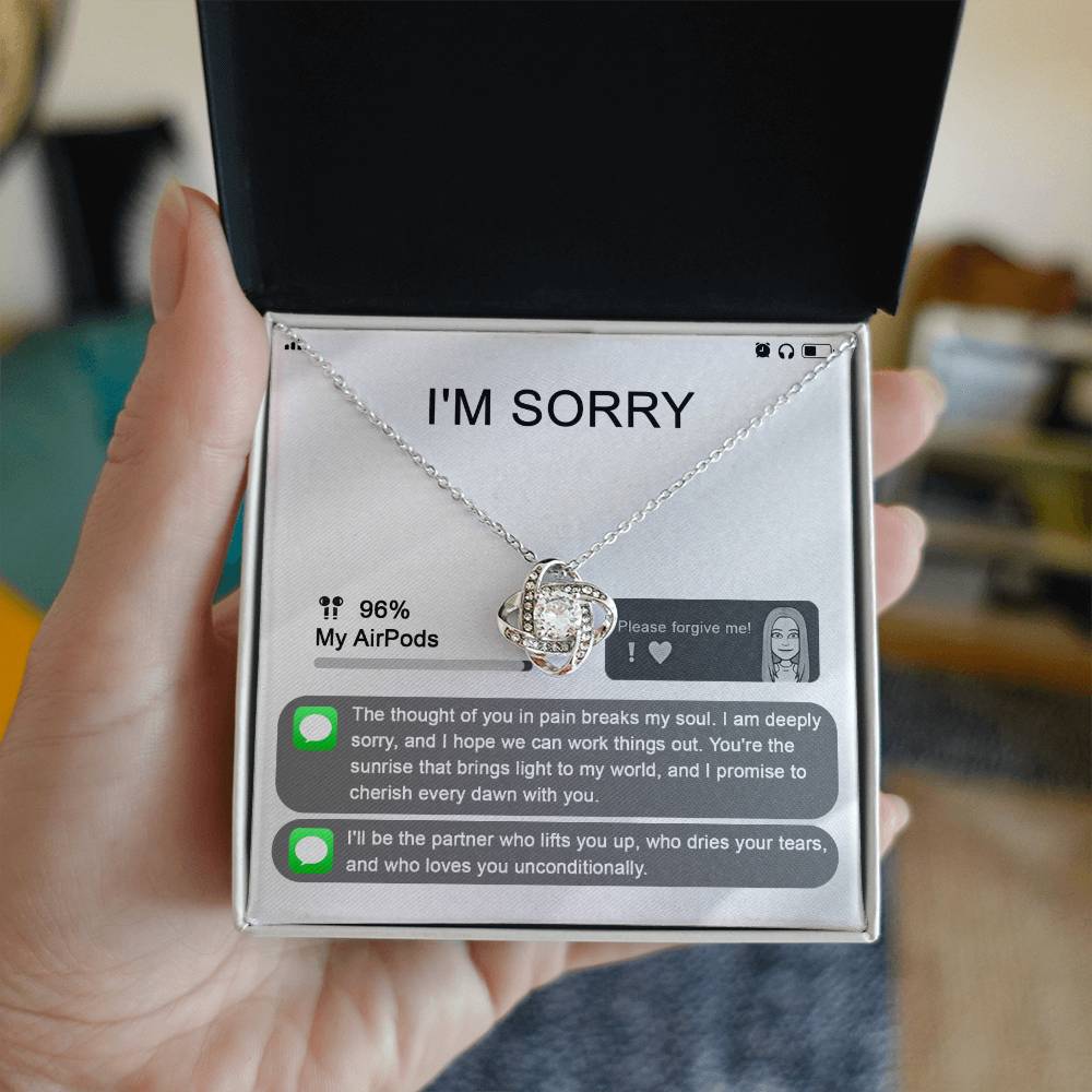 The "Sorry-Breaks My Soul - Love Knot Necklace" is a silver necklace featuring a pendant, beautifully displayed in its packaging. The front of the packaging includes a heartfelt apology message, text messages, a battery icon showing 96% for AirPods, and an illustrated figure. This intricately crafted piece is made from 14k white gold and adorned with cubic zirconia crystals.