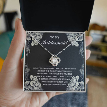 The "To Bridesmaid, The Luckiest Bride - Love Knot Necklace," adorned with cubic zirconia crystals, is elegantly presented on a gift box. The box comes with a heartfelt message expressing gratitude to a bridesmaid. Floral illustrations gracefully decorate the border, adding a charming touch to this piece available in either white gold or yellow gold finish.