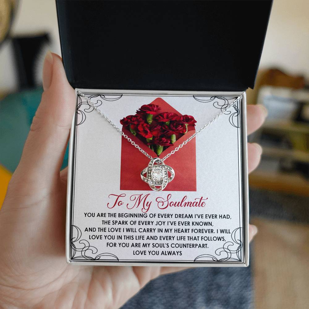 A hand displays an open jewelry box with the Soulmate-My Heart Forever - Love Knot Necklace, featuring a gold finish. Inside is a decorative card that reads "To My Soulmate," embellished with a romantic message and sparkling cubic zirconia accents.