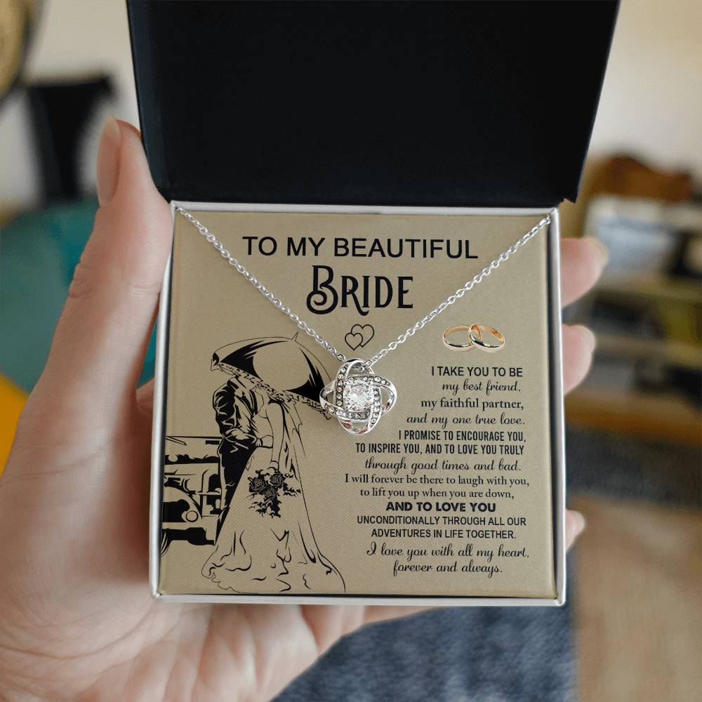 A "To My Bride, Faithful Partner - Love Knot Necklace" adorned with cubic zirconia crystals is displayed in a box. The background features a silhouette of a couple under an umbrella, two rings, and a heartfelt message to a bride, making it the perfect unique gift.