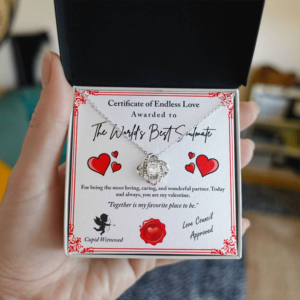 A hand presenting an open gift box unveils the exquisite Love Certificate-Cupid Witnessed - Love Knot Necklace, embellished with sparkling cubic zirconia crystals. The box lid features a certificate titled "The World's Best Soulmate," surrounded by red hearts, ideal for a personalized gift to your beloved.