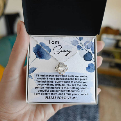 A hand holds an open box featuring the Sorry-Push You Away - Love Knot Necklace, adorned with cubic zirconia crystals and accompanied by an apology note that reads, "I am sorry...Please forgive me," embellished with blue floral designs.