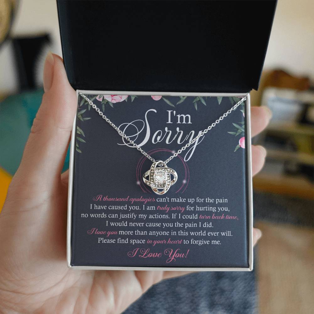 A box containing the "Sorry-World Ever Will - Love Knot Necklace," featuring a central pendant adorned with cubic zirconia crystals. The box lid displays text apologizing and seeking forgiveness, framed by a decorative floral border.