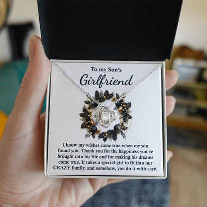 A hand holds a black box featuring the "Son_s Girlfriend- Found You - Love Knot Necklace," which has a circular pendant embellished with floral designs and enhanced with cubic zirconia. It conveys a message to "My Son's Girlfriend," expressing gratitude for her positive influence, making it an ideal personalized gift.