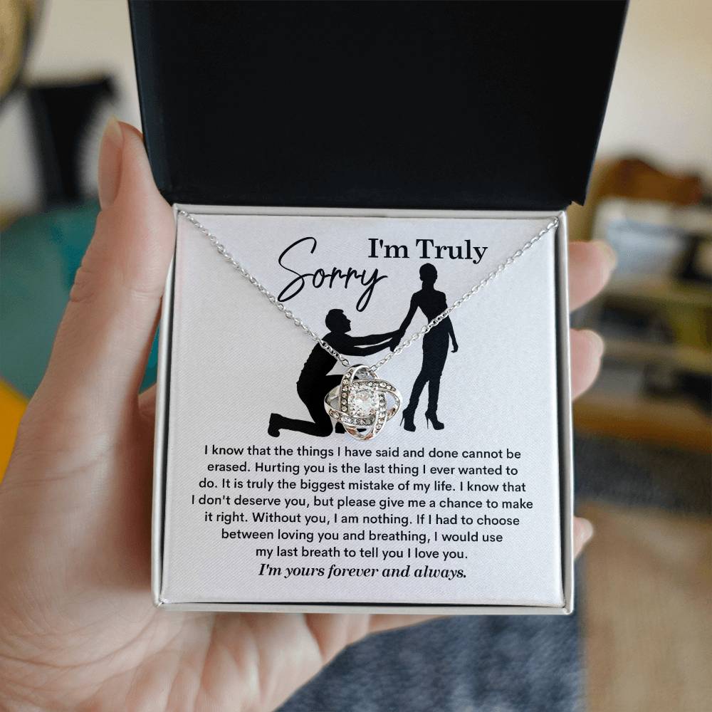 The Sorry-My Last Breath - Love Knot Necklace, a silver piece featuring an intertwined heart pendant decorated with cubic zirconia crystals, is presented on a card. The card includes an apology note with the phrase "I'm Truly Sorry" and the silhouette of one person kneeling before another.