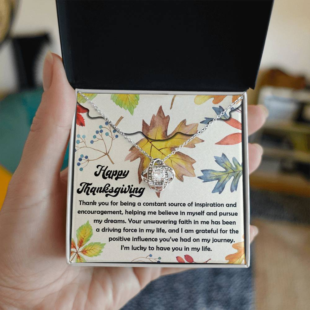 A hand holds an open gift box featuring the Thanksgiving-On My Journey - Love Knot Necklace, elegantly adorned with cubic zirconia. Behind the necklace, a card showcases a heartfelt Thanksgiving message alongside vibrant leaf illustrations, creating an ideal personalized gift.