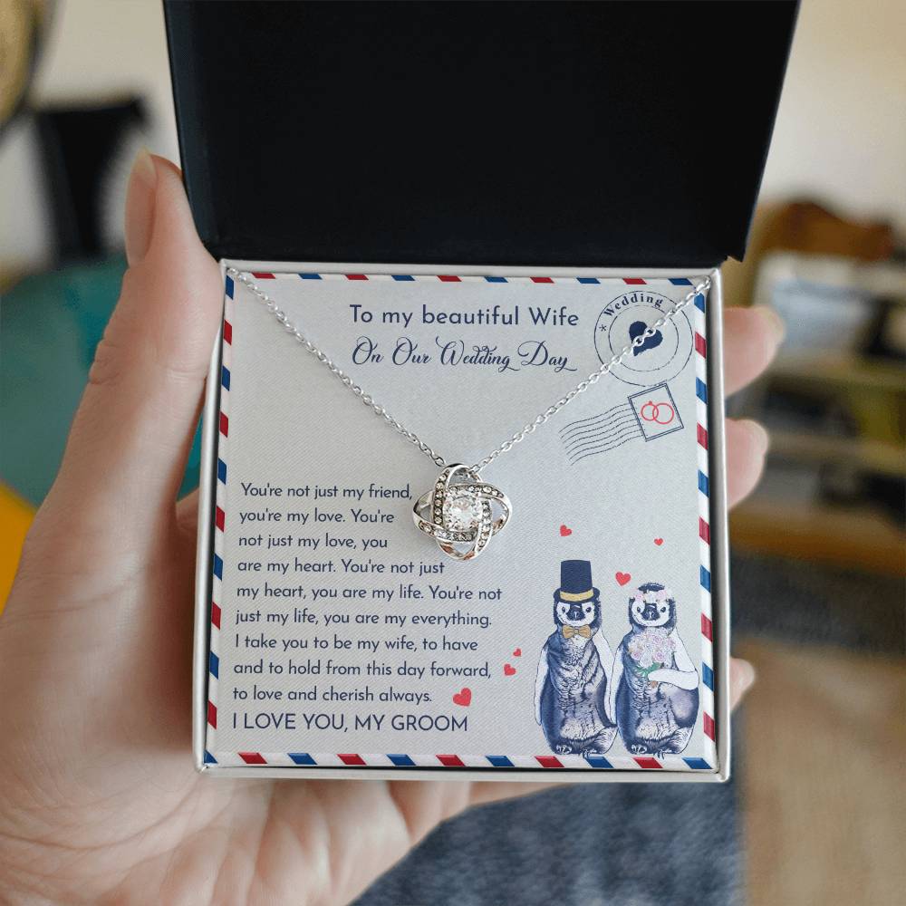 Close-up of a To Wife Wedding, Love And Cherish - Love Knot Necklace with cubic zirconia crystals on a card. The 14k white gold pendant is heart-shaped, and the card has a message for a wife on the wedding day. Two penguins, dressed as a bride and groom, are illustrated at the bottom.