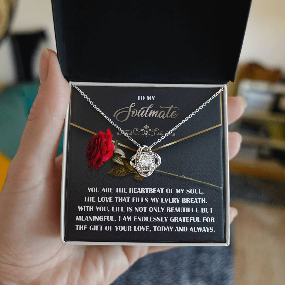 A hand holds an open jewelry box with the Soulmate-My Every Breath - Love Knot Necklace, featuring an ornate pendant and cubic zirconia sparkle, accompanied by a small red rose and a heartfelt message of gratitude and love for a soulmate—a truly personalized gift.