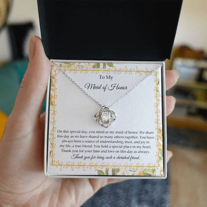 Presenting the "To Maid of Honor, Share This Day - Love Knot Necklace," featuring a stunning pendant adorned with cubic zirconia crystals and finished in white gold. It is beautifully displayed in a box embellished with a floral border and includes a heartfelt message expressing gratitude and appreciation to the "Maid of Honor.