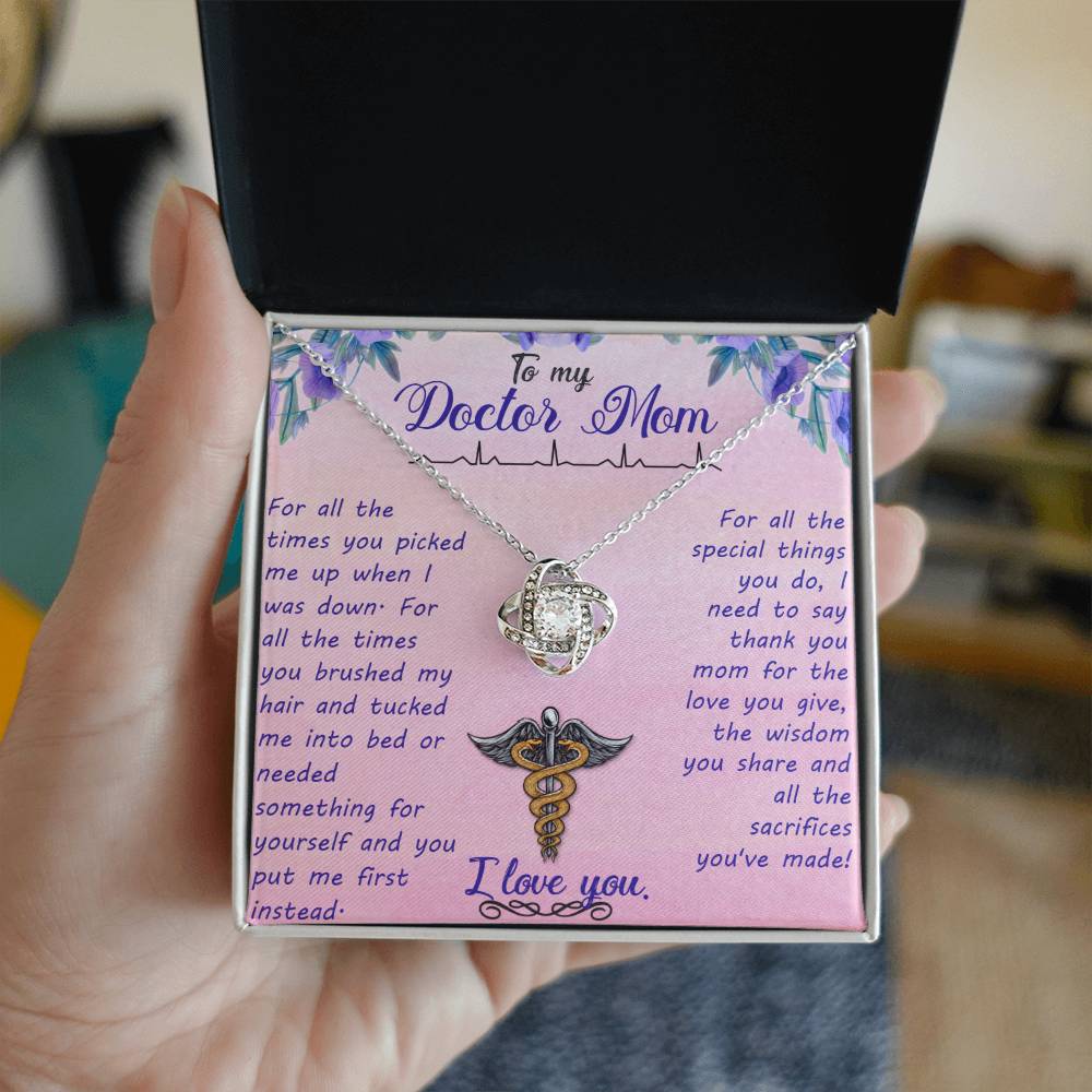 A hand holds an open gift box containing a ShineOn Fulfillment Love Knot Necklace with a medical symbol, displayed over a message card addressed to 'my doctor mom' expressing love and gratitude.