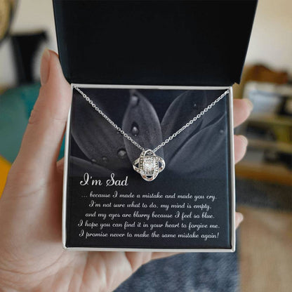 The "Sorry-Feel So Blue - Love Knot Necklace," featuring a stunning knot pendant adorned with cubic zirconia crystals, is displayed over a card with an apologetic message starting with "I'm Sad" and set against a floral background. This personalized gift beautifully combines elegance and heartfelt sentiment.