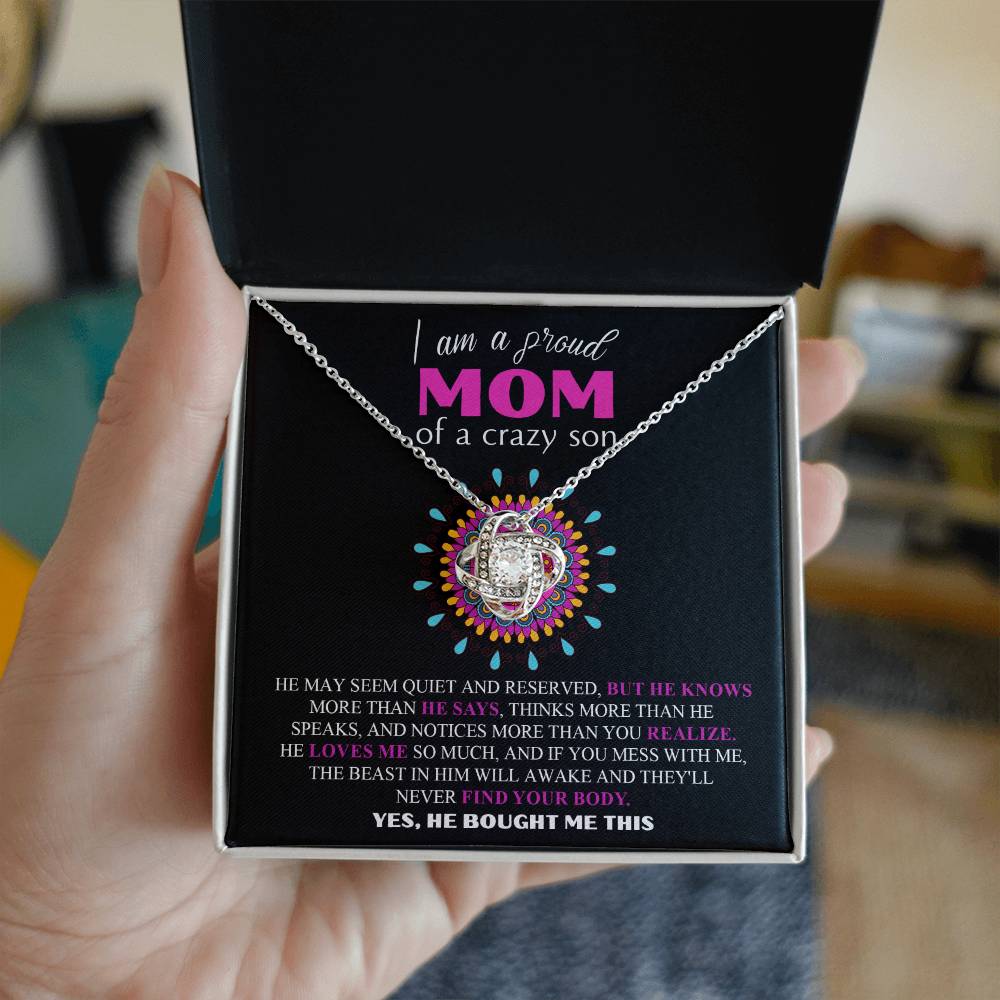 A person's hand holding the stunning "To Mom, Proud Mom - Love Knot Necklace" adorned with cubic zirconia crystals, with a background message that reads: "I am a proud MOM of a crazy son..." displayed in a black box. The necklace features an intricately decorated pendant.