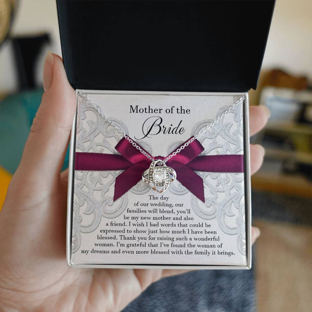 A To Bride's Mom, My New Mother - Love Knot Necklace with a heart pendant, crafted from 14k white gold and adorned with cubic zirconia crystals, is displayed on a decorative card with a maroon ribbon. The card reads, "Mother of the Bride" and contains a heartfelt message of gratitude.