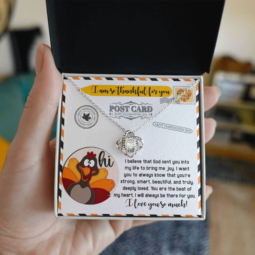 A hand holds an open box displaying the Thanksgiving-The Beat - Love Knot Necklace above a Thanksgiving card adorned with a turkey illustration and a heartfelt message. Inside the box, the necklace with its gold finish glistens, and its cubic zirconia sparkles like autumn leaves in the sunlight.