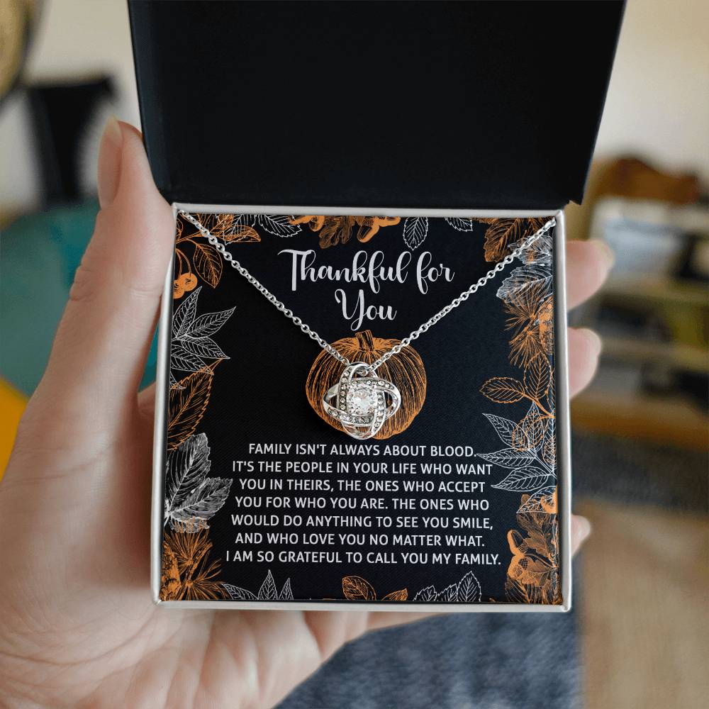 A person holds an open box revealing the stunning Thanksgiving-To Call You - Love Knot Necklace, elegantly finished in white gold. Accompanying it is a heartfelt card adorned with a sentimental family message, beautifully highlighted by shimmering cubic zirconia accents.